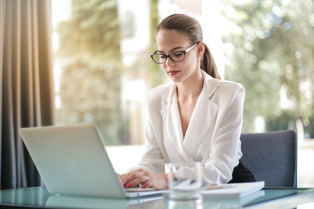 vrouw met witte blouse, bril en staart achter laptop aan het werk