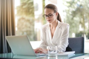vrouw met witte blouse, bril en staart achter laptop aan het werk